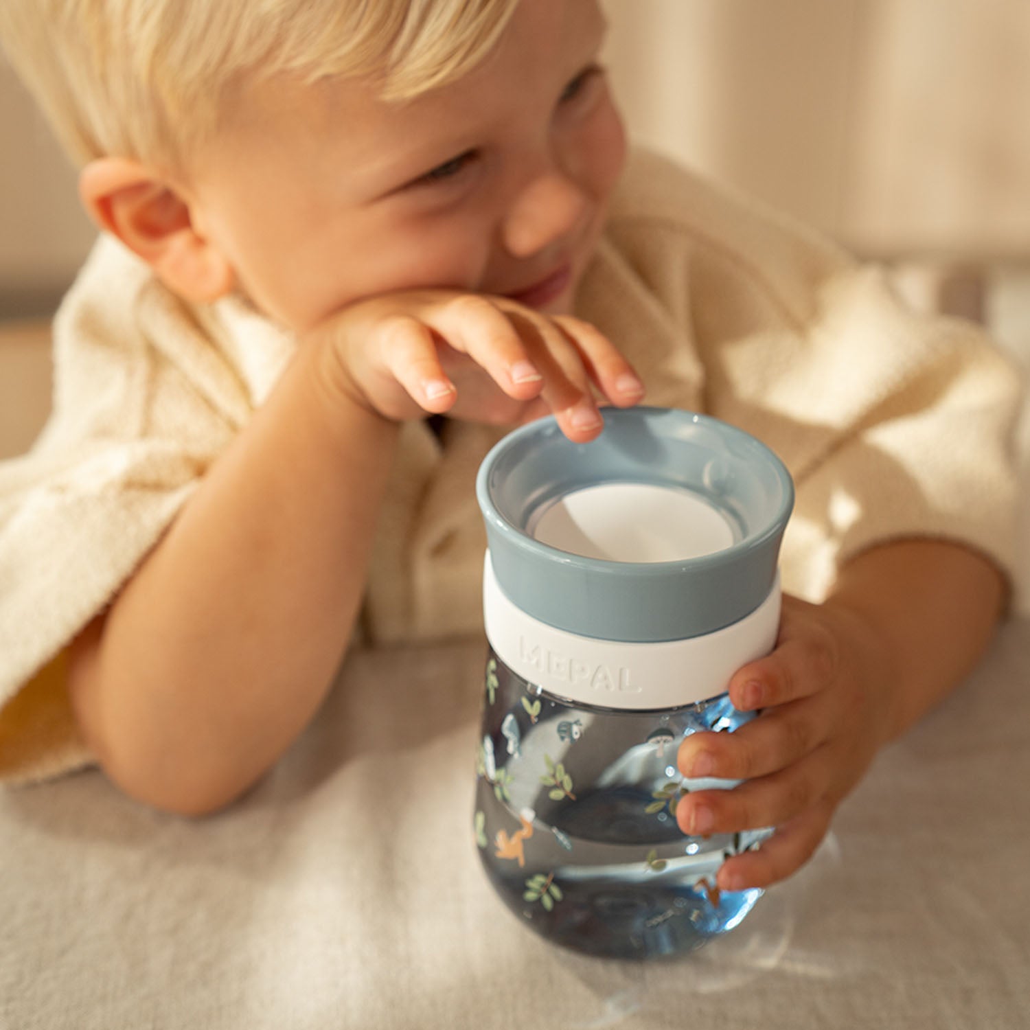 A smiling young boy resting his hand on the lid of the Little Dutch 360-degree Forest Friends cup on a table, highlighting its child-friendly, spill-resistant design.