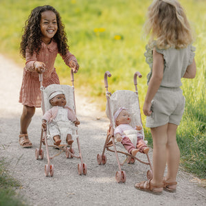 Two young girls playing outdoors on a dirt path, pushing their Little Dutch pink doll strollers.