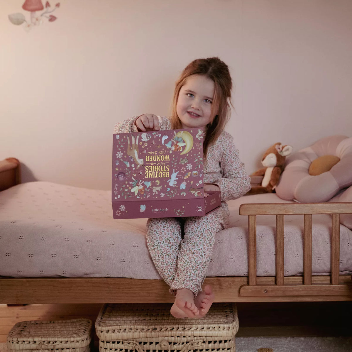 A young girl with light brown hair sits on a wooden bed holding a decorative Little Dutch pink story projector flashlight box. She is wearing a light pink and white patterned pajama set.