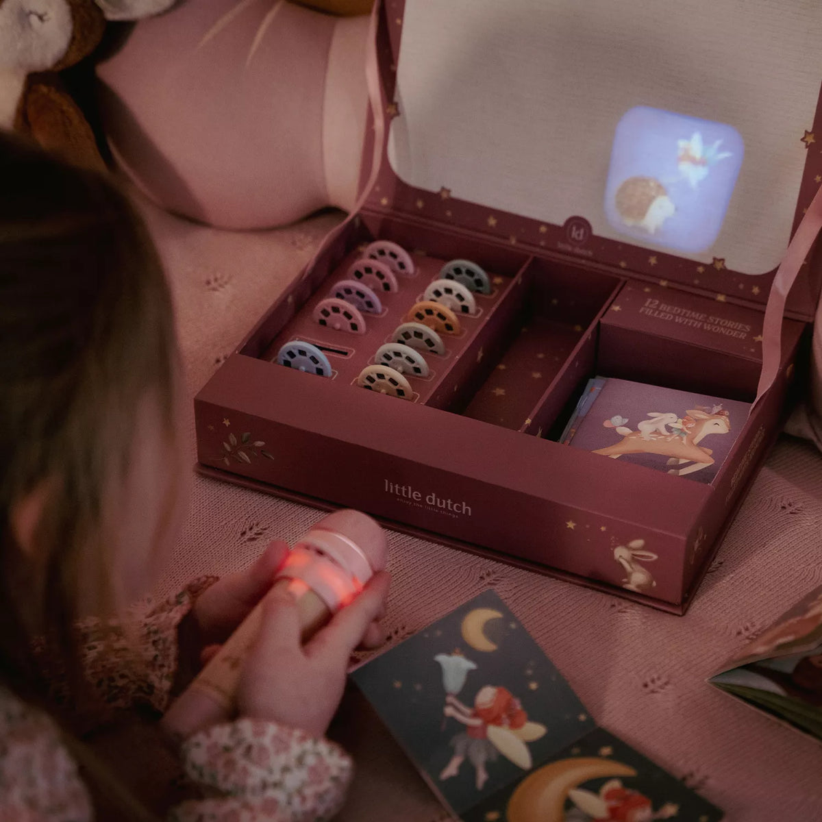 A child holds a small glowing fairy light in a cozy bedroom setting, with an open 'little dutch story projector flashlight gift box showing a set of colorful discs and booklet  on a pink bedspread.