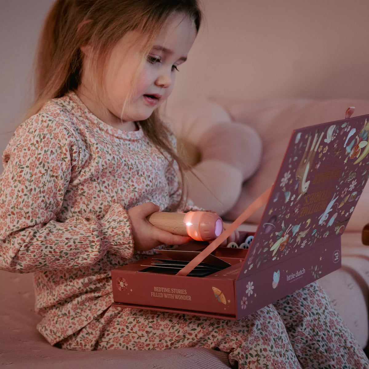 A young child in floral pajamas sits on a couch holding a flashlight over an open little dutch story projector box. The scene is softly lit and cozy, with the child as the main subject in a real indoor environment.