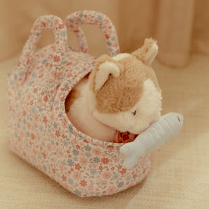 The plush cat sitting inside its floral carry bag while holding the blue toy fish in its mouth.