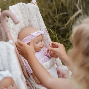 Close-up of a child's hands adjusting the pink headband on the Little Dutch Baby Lily doll, which is lying in a woven basket.