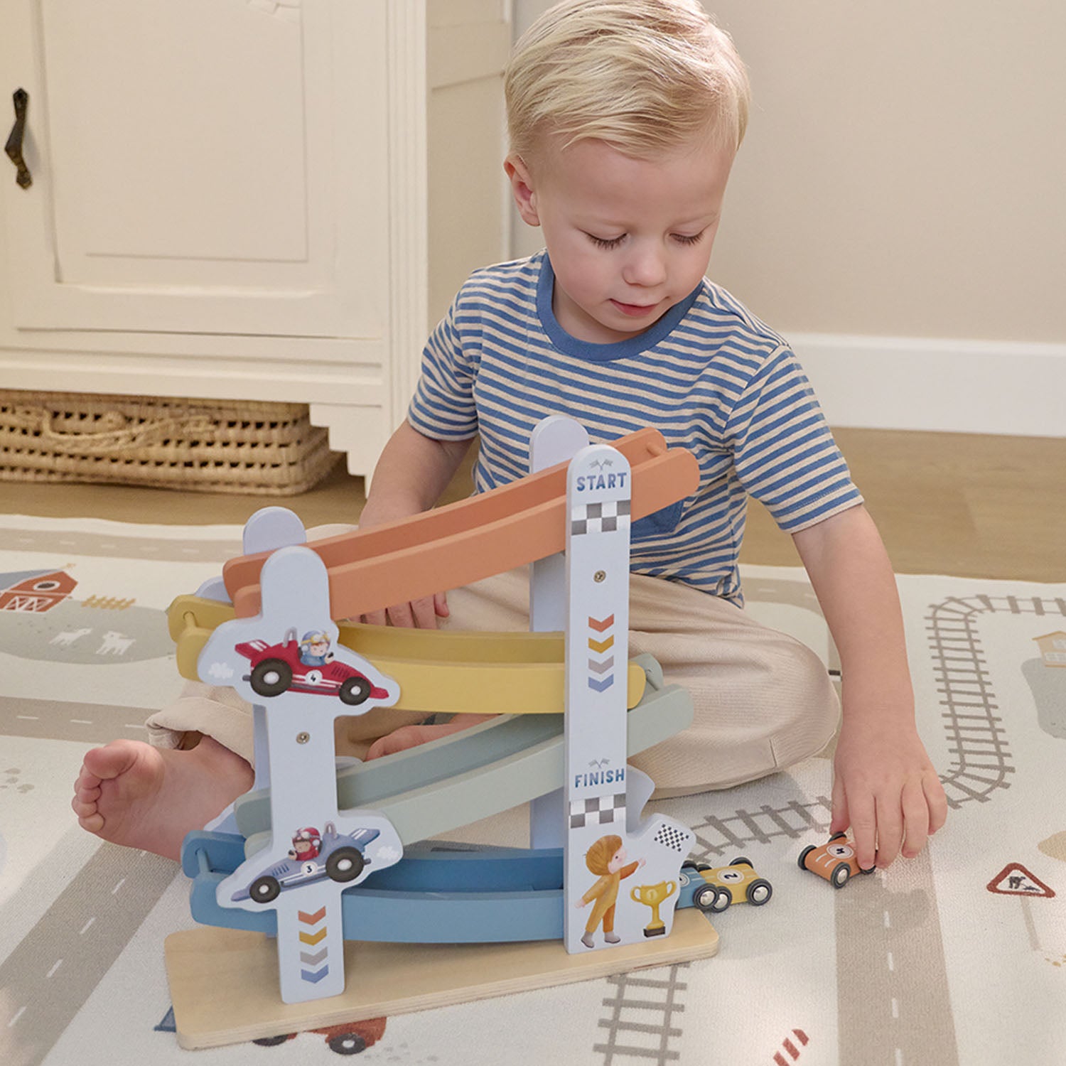 A young boy sitting on the floor, playing with the blue and orange transport-themed wooden ramp racer toy.