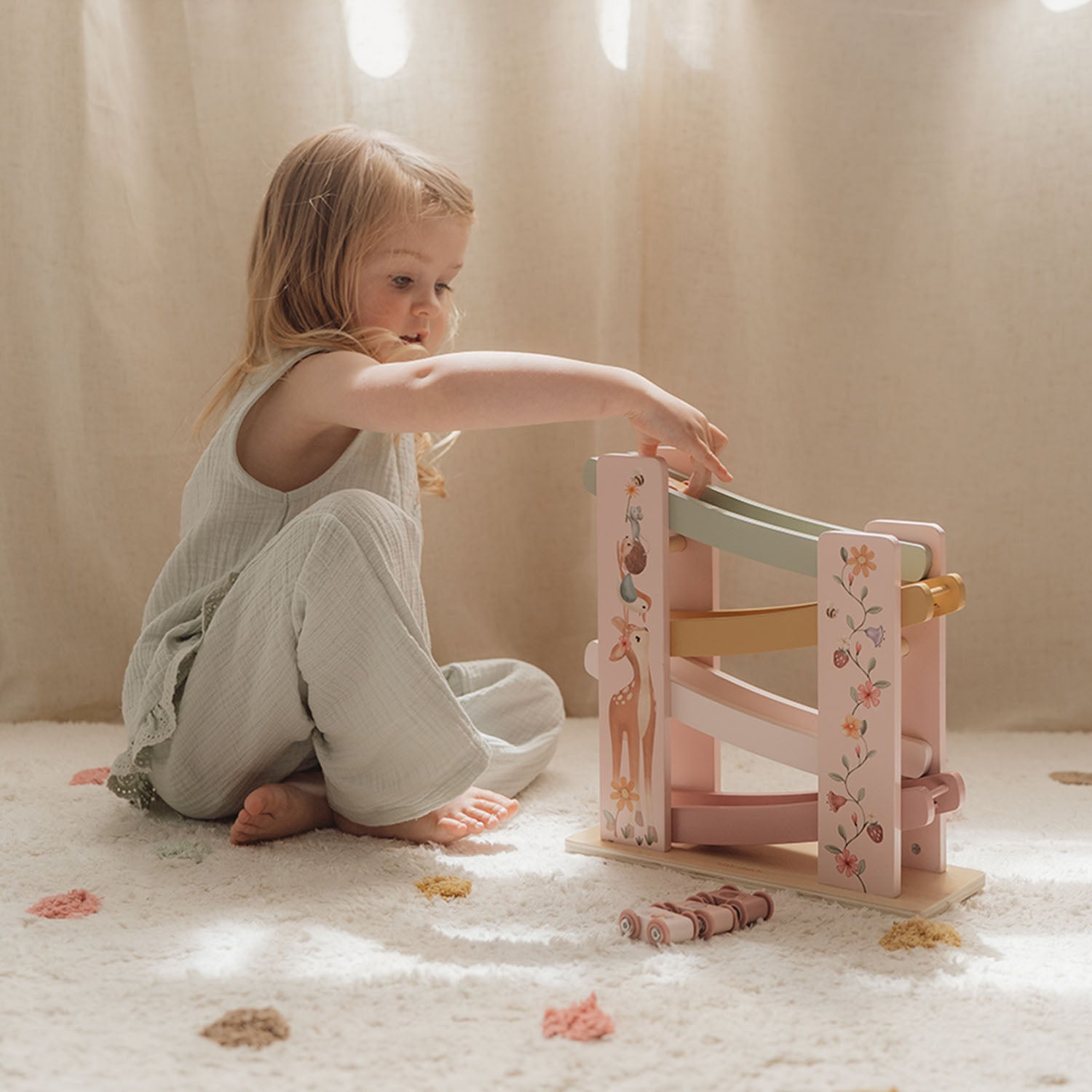 A young girl sitting on the floor, actively playing with the pink floral wooden ramp racer toy.