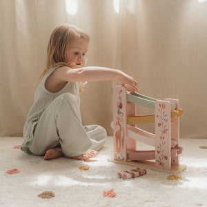 A young girl sitting on the floor, actively playing with the pink floral wooden ramp racer toy.