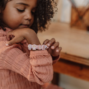 A close-up lifestyle shot of a young child wearing a pastel pink and white lettered bracelet on their wrist, wearing a textured dusty rose long-sleeve top.