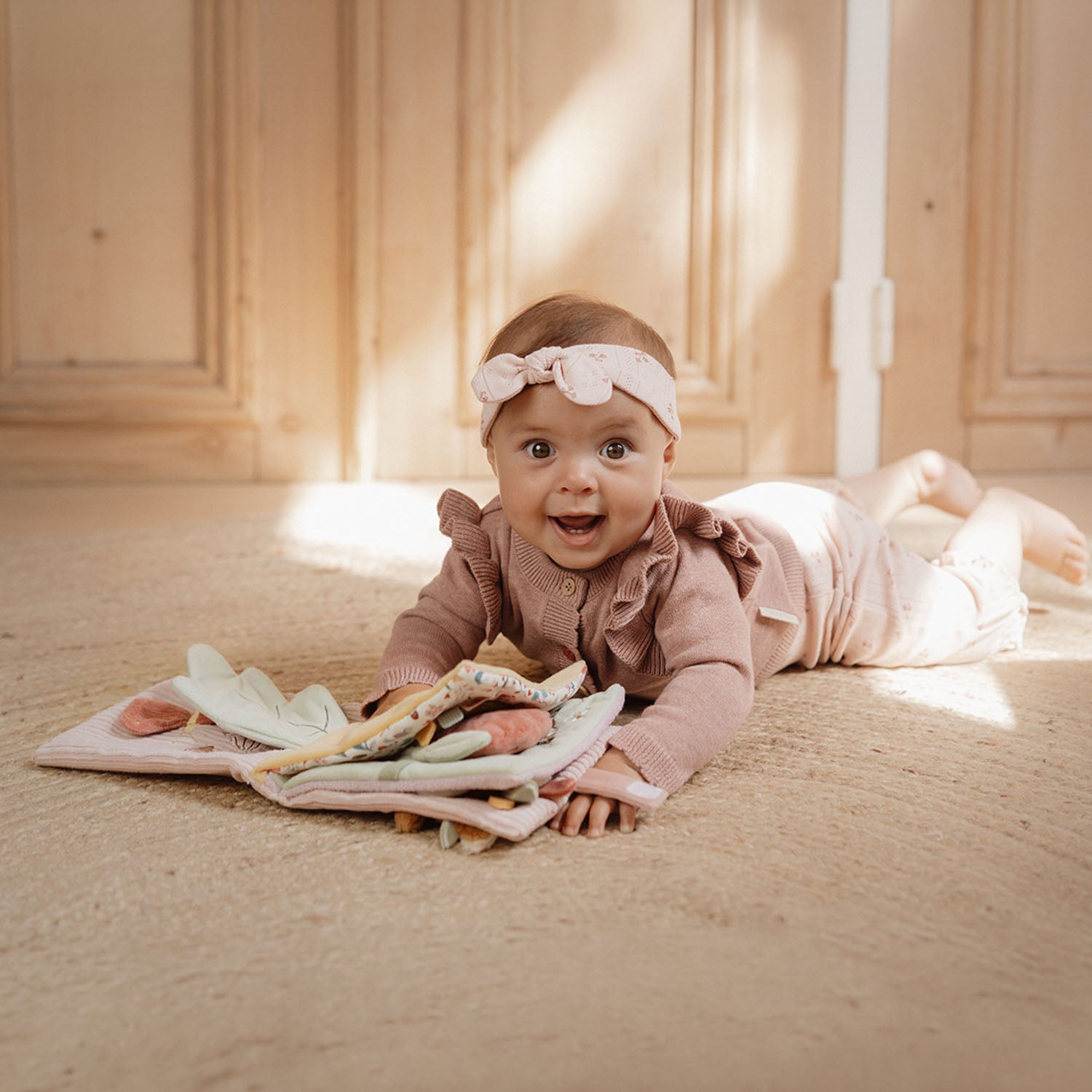 Baby lying on a wooden floor with toys, wearing a pink outfit and headband.