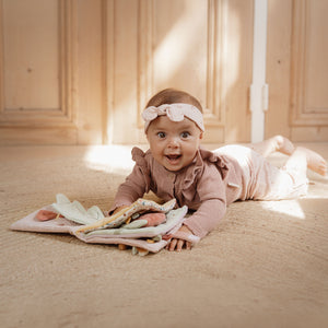 Baby lying on a wooden floor with toys, wearing a pink outfit and headband.