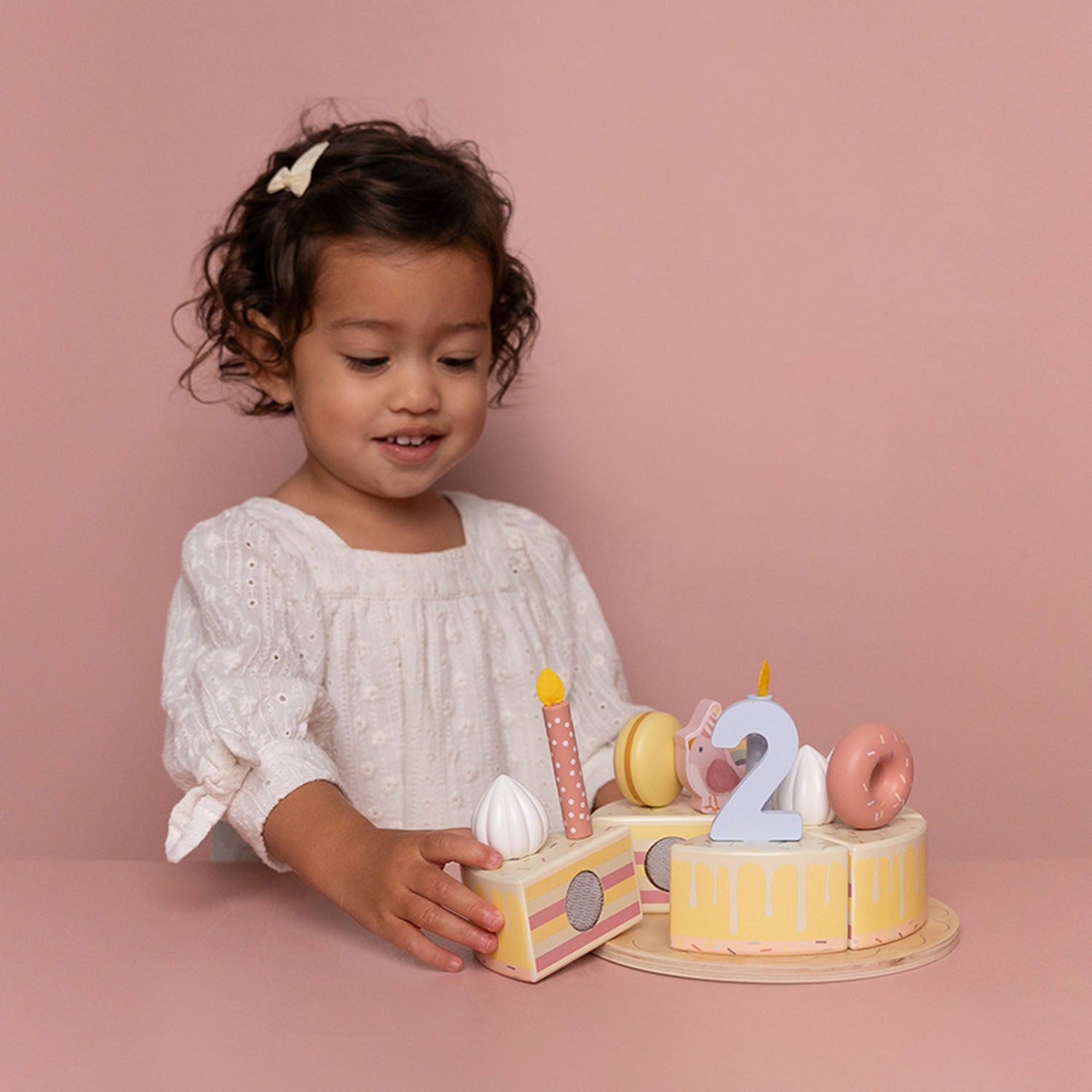 A smiling young girl sitting and playing with the pink Little Dutch wooden birthday cake play set on a pink background.