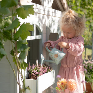 Child in a pink dress watering plants with a toy watering can in a garden setting.