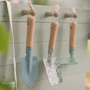 Close-up of small blue and patterned garden hand tools hanging from wooden pegs on the backboard of a mud kitchen.