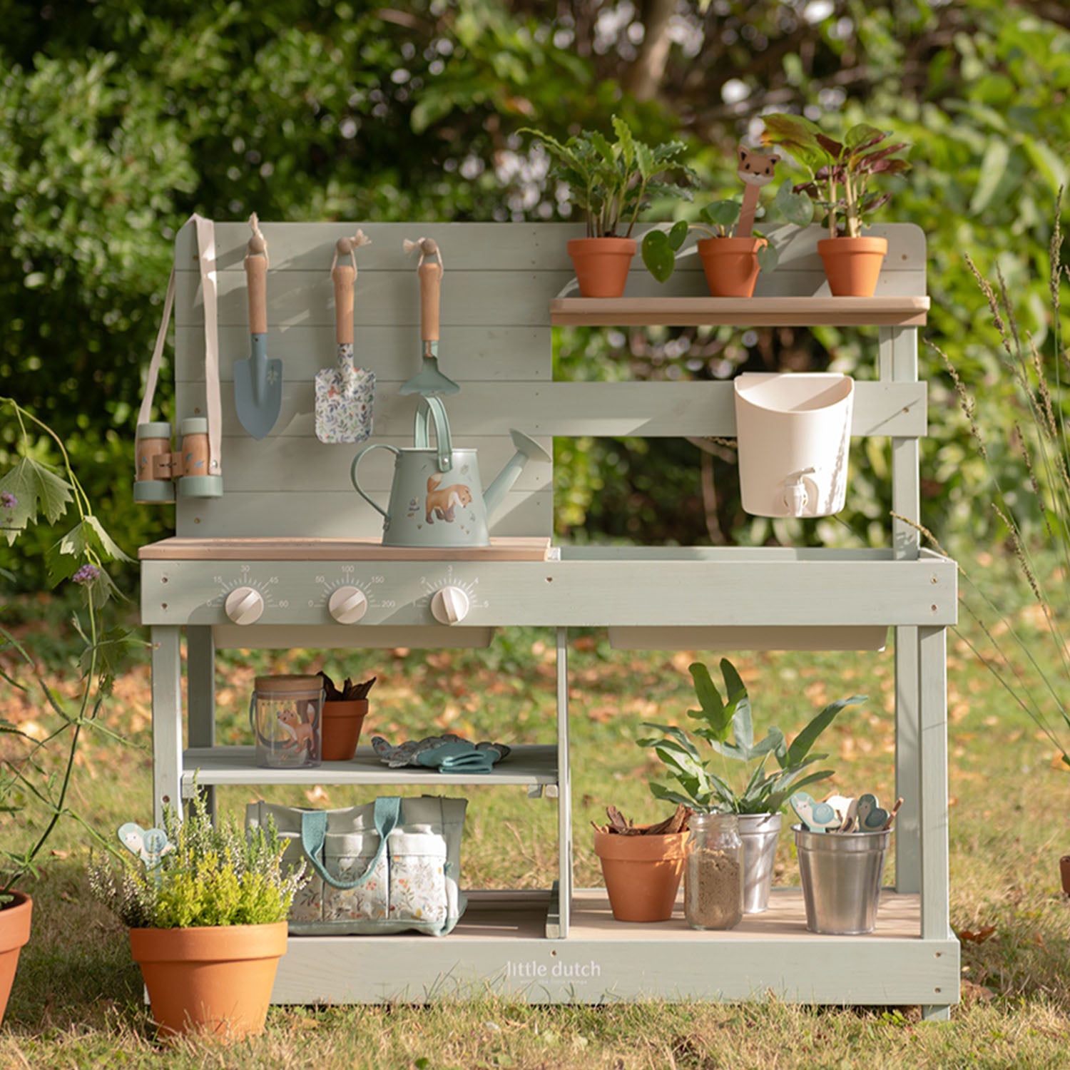 Little Dutch wooden mud kitchen fully styled with potted plants and garden tools outdoors on a sunny lawn.