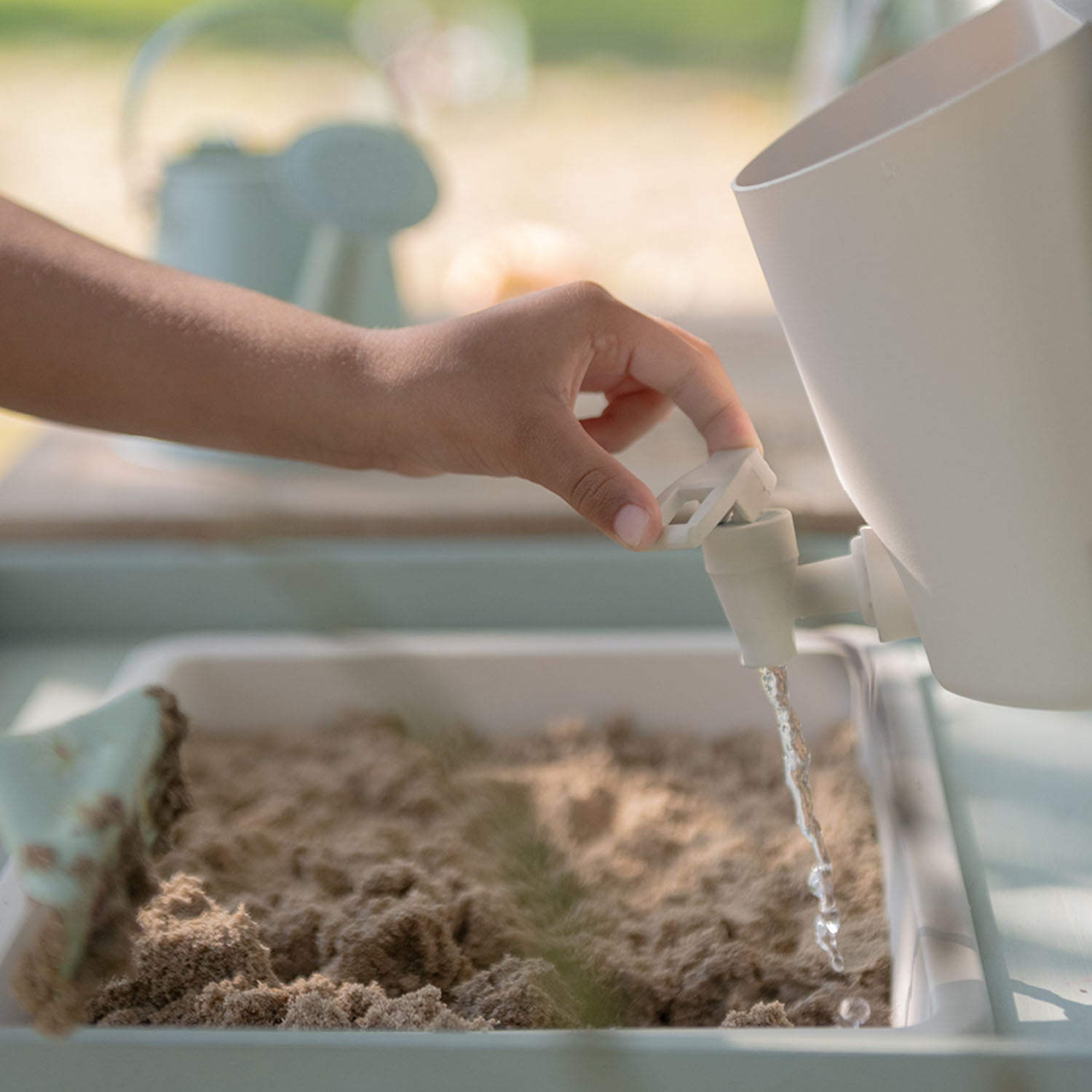 Close-up of a child using the working water tap and reservoir to pour water into a basin filled with sand.