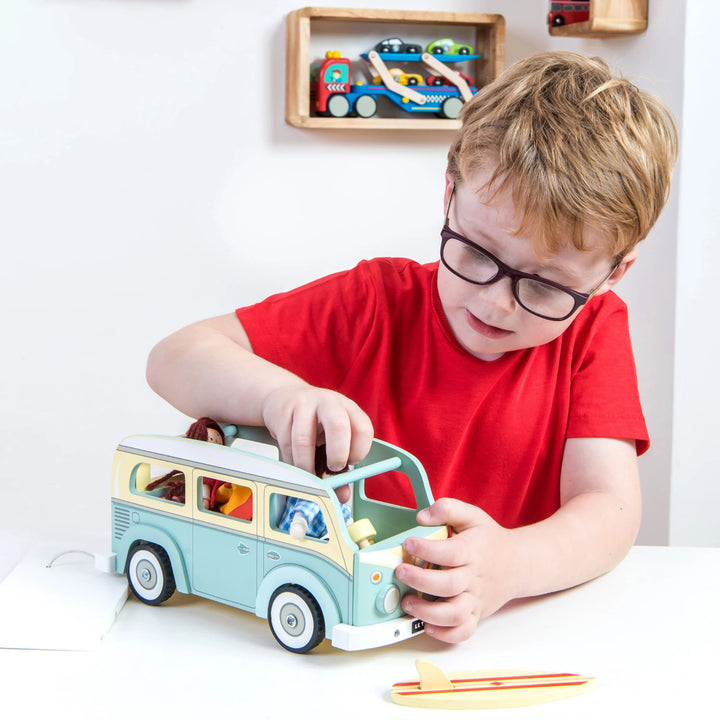 A child in a red shirt playing with a wooden Le Toy Van camper van. The van is blue and white with a red surfboard on top