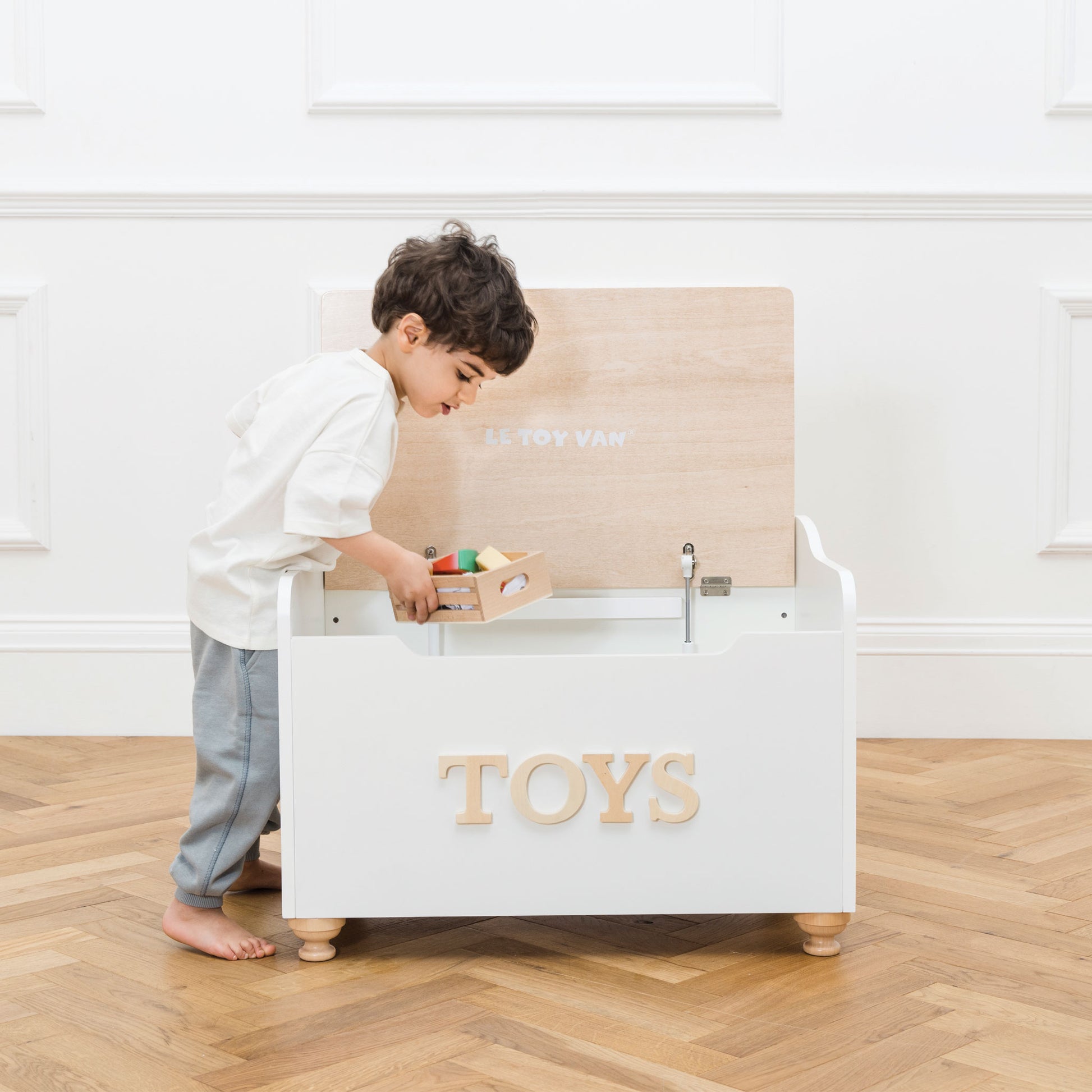 Child playing with a toy box labeled 'TOYS' on a wooden floor.