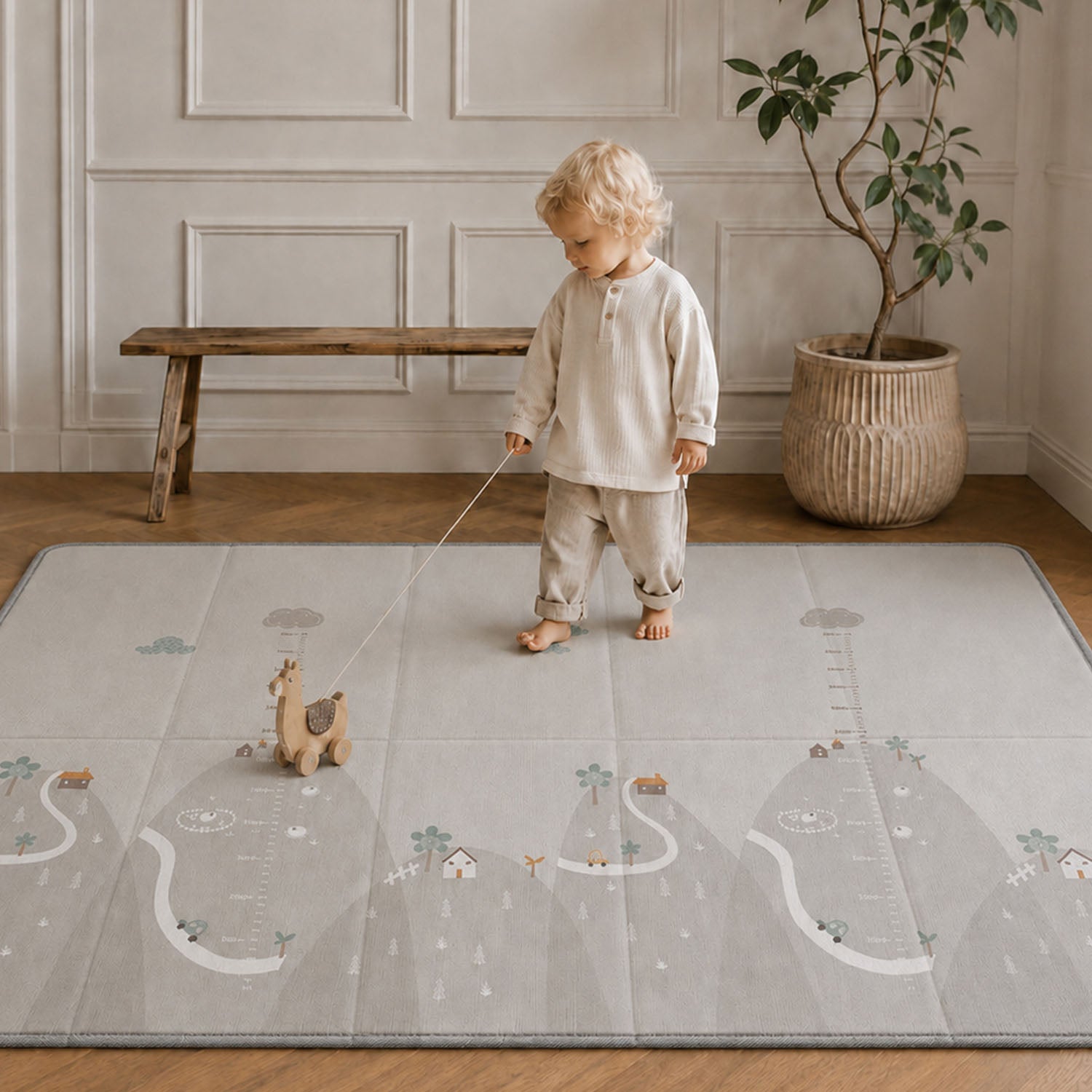 A toddler stands barefoot on a large light gray play mat with a whimsical landscape print, holding a string attached to a small toy. The room has wood flooring, a wooden bench, and a potted plant in the background.