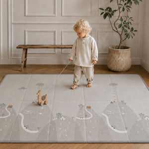 A toddler stands barefoot on a large light gray play mat with a whimsical landscape print, holding a string attached to a small toy. The room has wood flooring, a wooden bench, and a potted plant in the background.