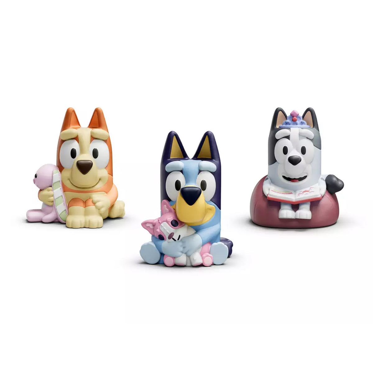 Tonies Three Bluey-inspired dog character figurines posed together on a clean white background: a yellow-and-orange dog holding a pink plush, a blue-and-white dog holding a pink cat, and a gray-and-white dog sitting on a burgundy cushion with a book.