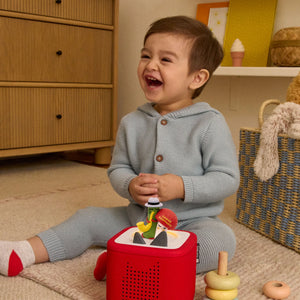 A smiling toddler sitting on the floor, interacting with the Caspar Babypants Tonie figure placed on top of a red Toniebox.