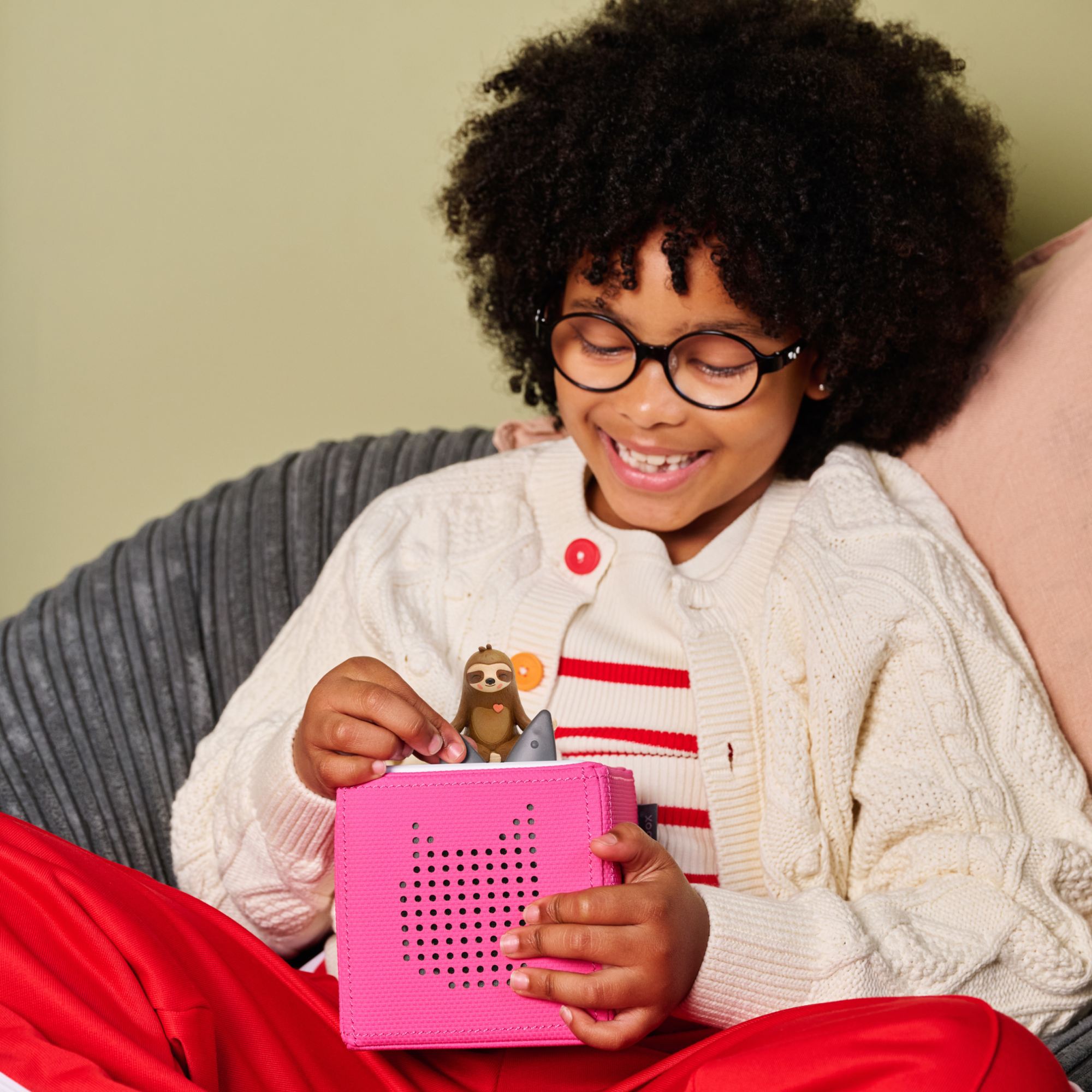 A smiling child with curly hair and glasses playing with the Mo the Sloth Tonie and a pink Toniebox.