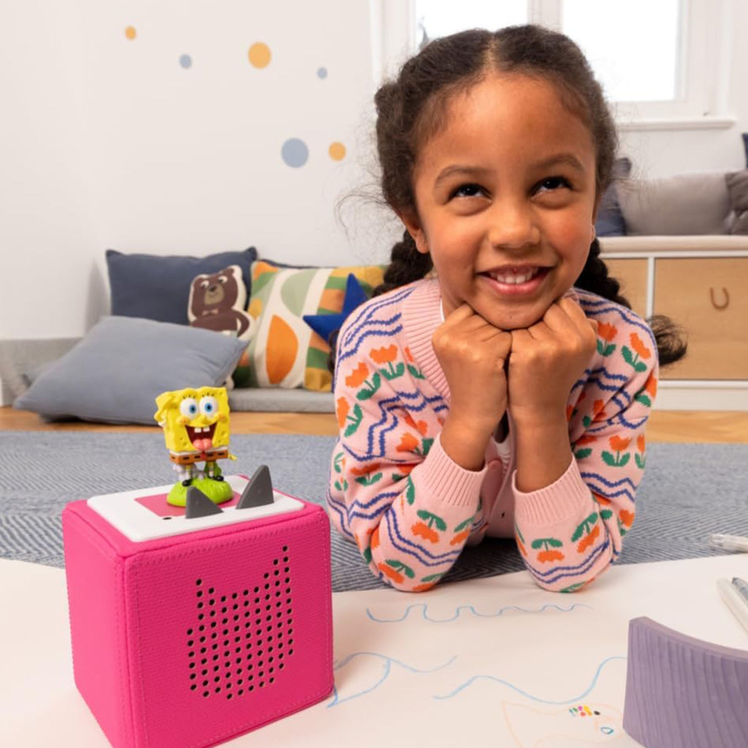 A smiling young girl happily listening to the SpongeBob SquarePants Tonie playing on a pink Toniebox.