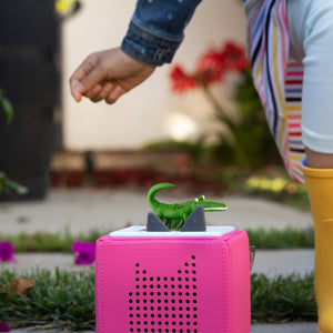 A child's hand placing the green Enormous Crocodile Tonie figurine onto the top of a pink Toniebox speaker outdoors.