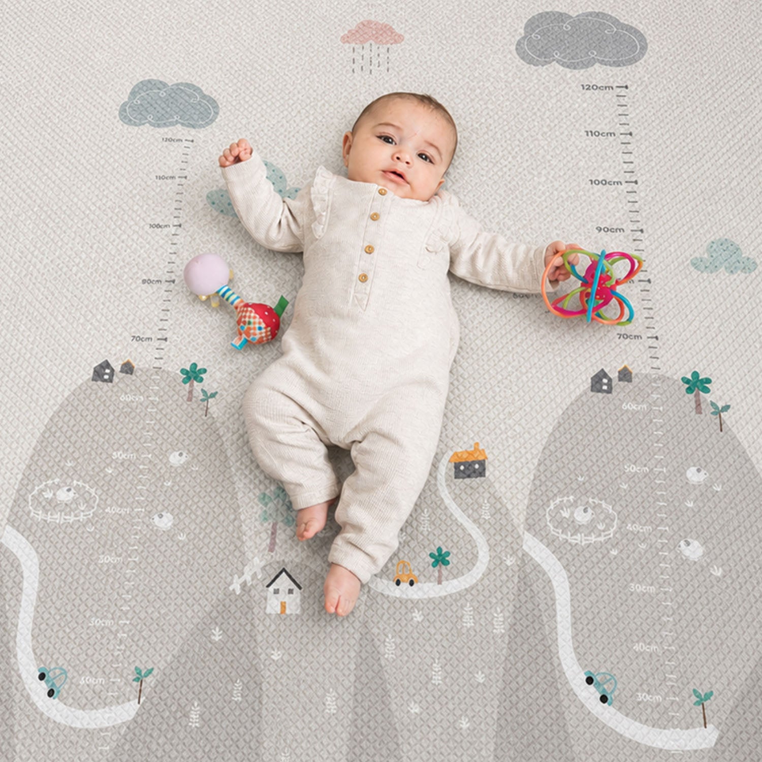 A baby lies on a printed baby growth chart mat with cloud, house, and tree illustrations. The baby is wearing a cream-colored long-sleeve romper and holding a colorful rattle.