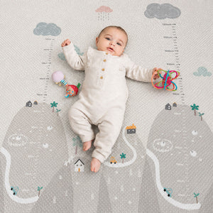 A baby lies on a printed baby growth chart mat with cloud, house, and tree illustrations. The baby is wearing a cream-colored long-sleeve romper and holding a colorful rattle.