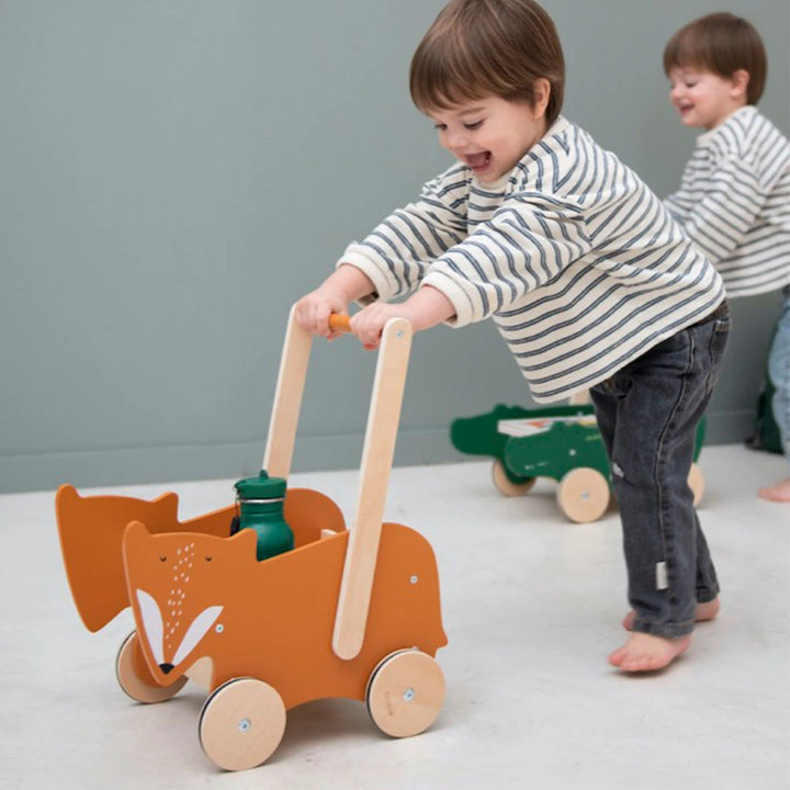 A toddler pushing the Trixie Wooden Push Along Cart - Mr. Fox filled with toys. The cart is made of wood and painted with colorful fox character on the front.