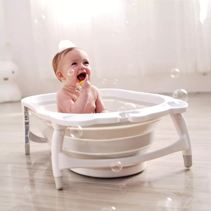 A baby happily sitting in a blue and white Karibu foldable baby bath tub placed on a wooden floor, with bubbles around indicating bath time.