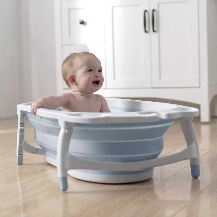 A joyful baby sitting in a grey Karibu foldable baby bath tub, playing with a yellow bath toy and surrounded by floating soap bubbles.