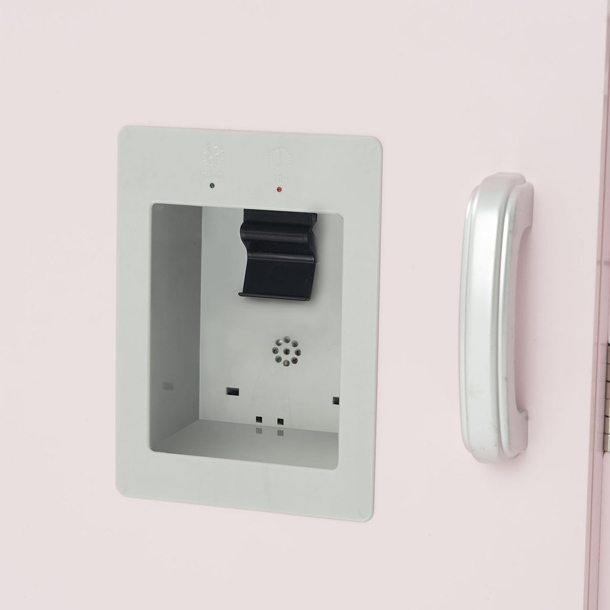 A close-up of a light pink toy kitchen refrigerator door showing the water dispenser compartment with a black drip tray and a white curved handle.