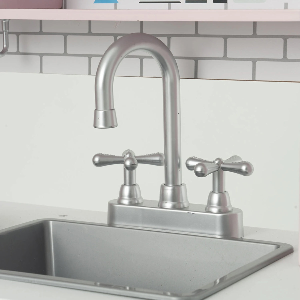 A brushed silver double-handle play kitchen faucet with a high-arc spout, installed in a stainless steel sink against a white subway tile backsplash.