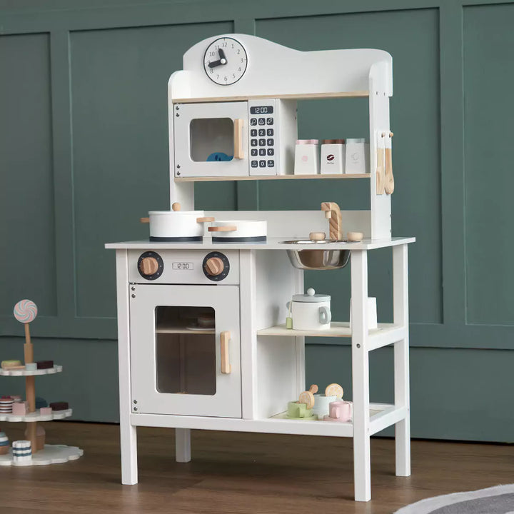 A small, white wooden play kitchen with an oven, stovetop, microwave, sink, and storage shelves. It also includes five toy utensils.