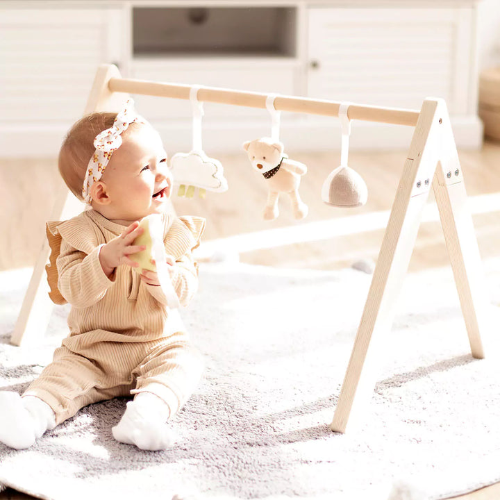 Baby smiling and reaching for colorful hanging toys on a wooden play gym with a green leaf playmat.